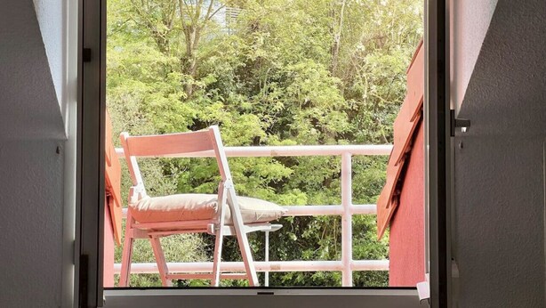 A balcony with a wooden chair and a table, overlooking a garden with trees and a building in the background. - El Solsticio - 30601 (Somo)