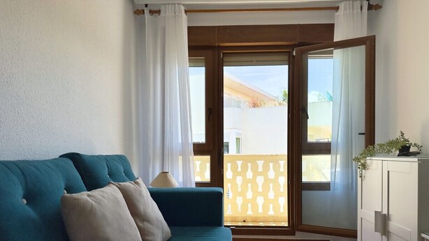 Ein Zimmer mit einem türkisfarbenen Sofa, einem weißen Schrank und Blick durch eine Glastür auf einen Außenbereich mit einem gemusterten Zaun.