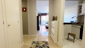 A hallway with a kitchen area on the right, a small stool, and a door with a red emergency light.