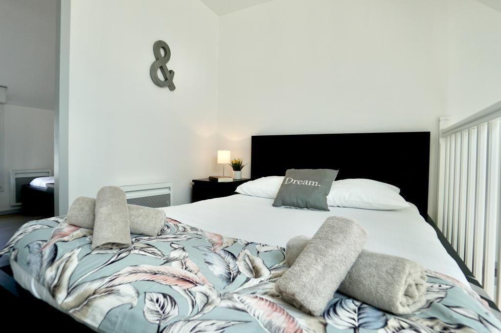 Un lit bien fait, recouvert d’une couvre-lit à motifs floraux, un oreiller orné du mot « Rêve », et un luminaire fixé au mur.