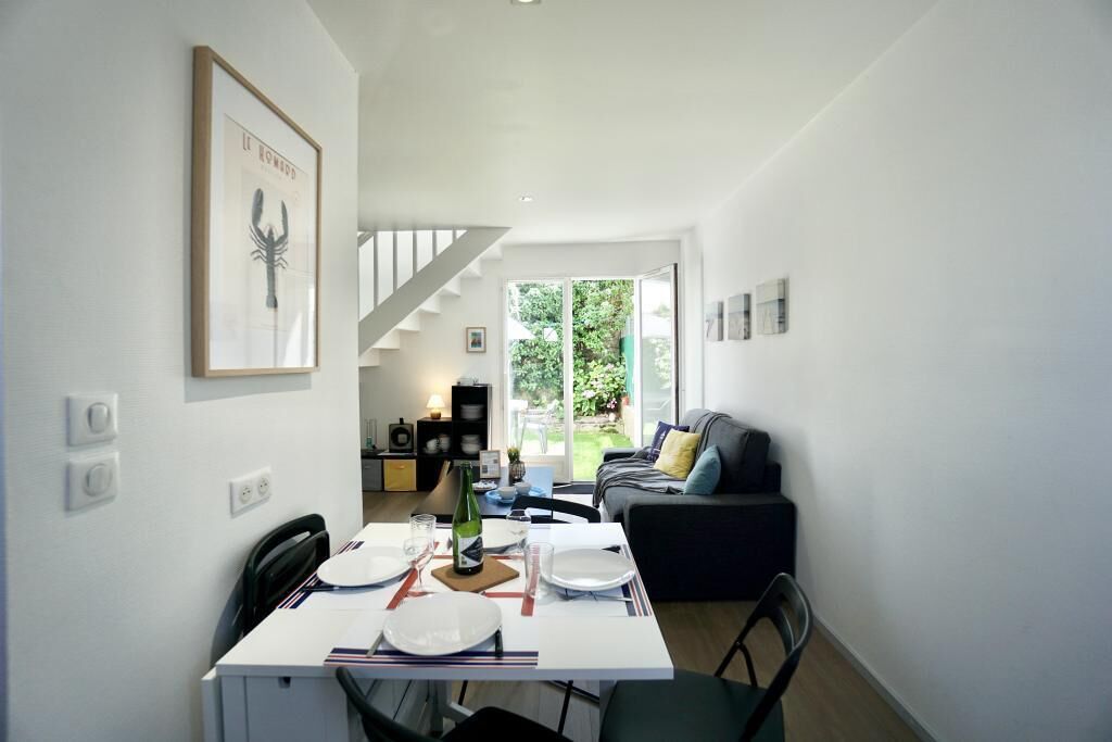 A modern dining area with a white table set for two, a staircase leading to a balcony, and a dark-colored sofa in the background.