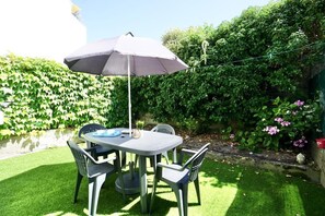 An outdoor patio with a table and chairs, a grey umbrella, and a hedge in the background.