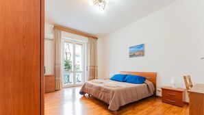 A bedroom with a wooden floor, a bed with a blue pillow, a nightstand, a dresser, and a window with curtains. - 'Dante House' Apartment in the centre ideal for groups (Cagliari)