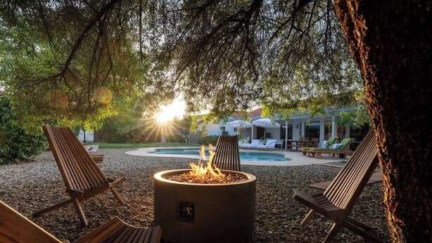 A fire pit surrounded by wooden lounge chairs in a landscaped outdoor area with a pool and umbrellas in the background. - Sleeps 18 | Beautiful Resort Style | Heated Pool (Scottsdale)