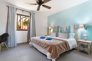 Een slaapkamer met een bed, gordijnen, een plafondventilator, een nachtkastje met lamp en een stoel.
