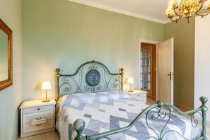 Une chambre à coucher comprenant un lit, deux tables de chevet, une commode, un lustre et une porte donnant sur une autre pièce.