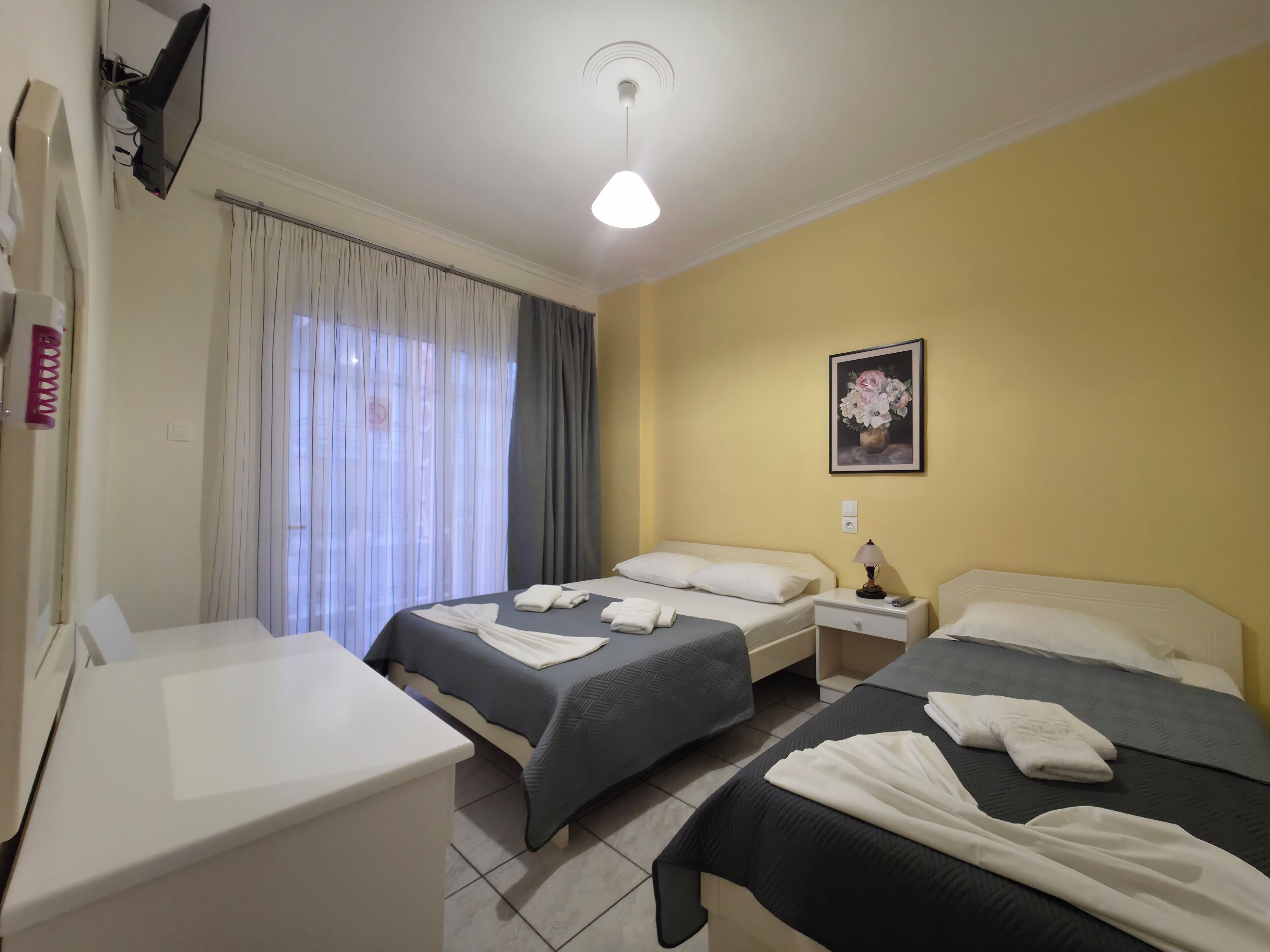 A hotel room with two beds, a bedside table with a lamp, a large window with curtains, and a painting on the wall.