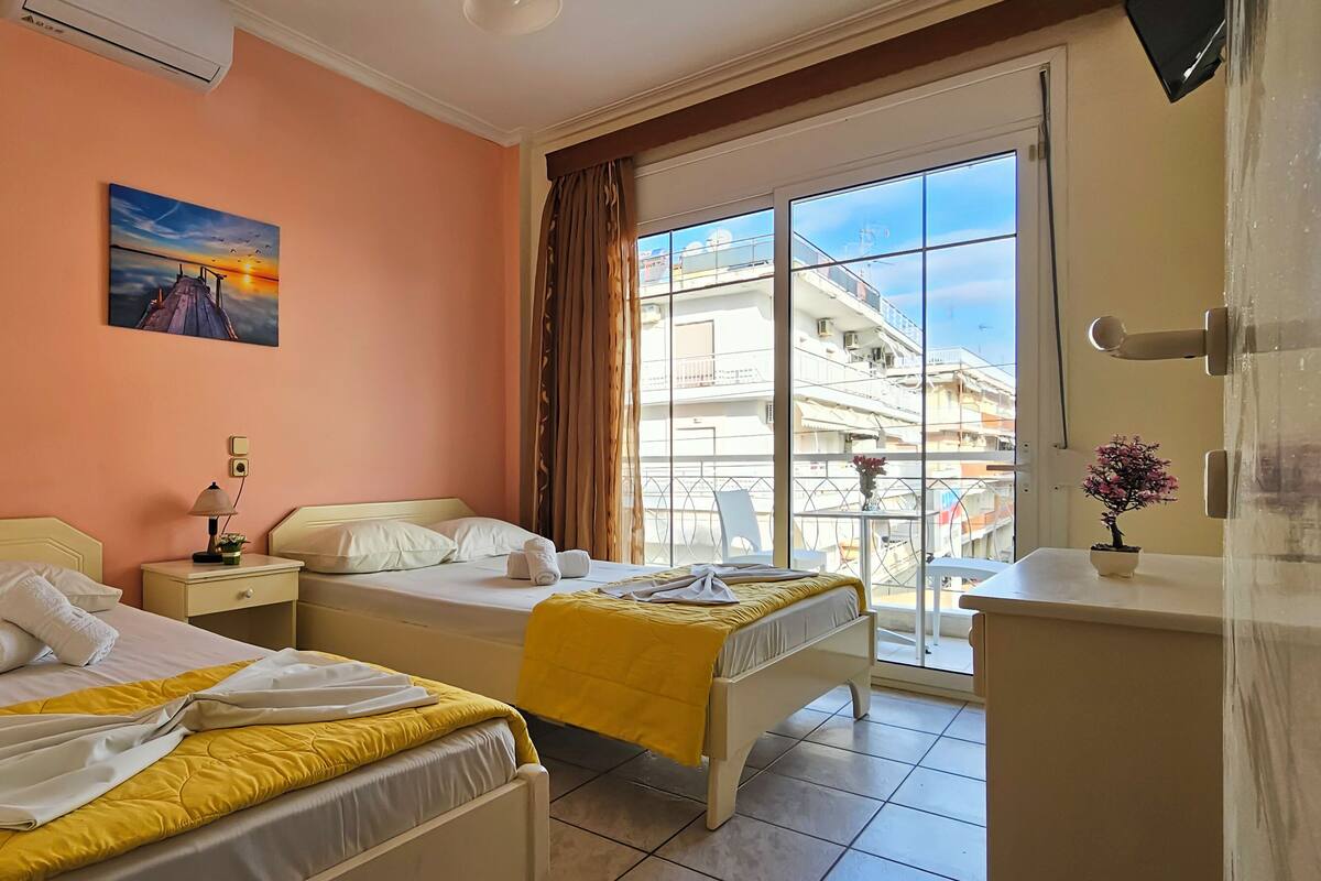 A hotel room with two beds, a window with a view of buildings, a painting on the wall, and a small table with a plant.