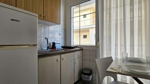 A compact kitchen with wooden cabinets, a white refrigerator, a window with sheer curtains, and a dining area with a table set for two. - Chariclia Hotel (Katerini)