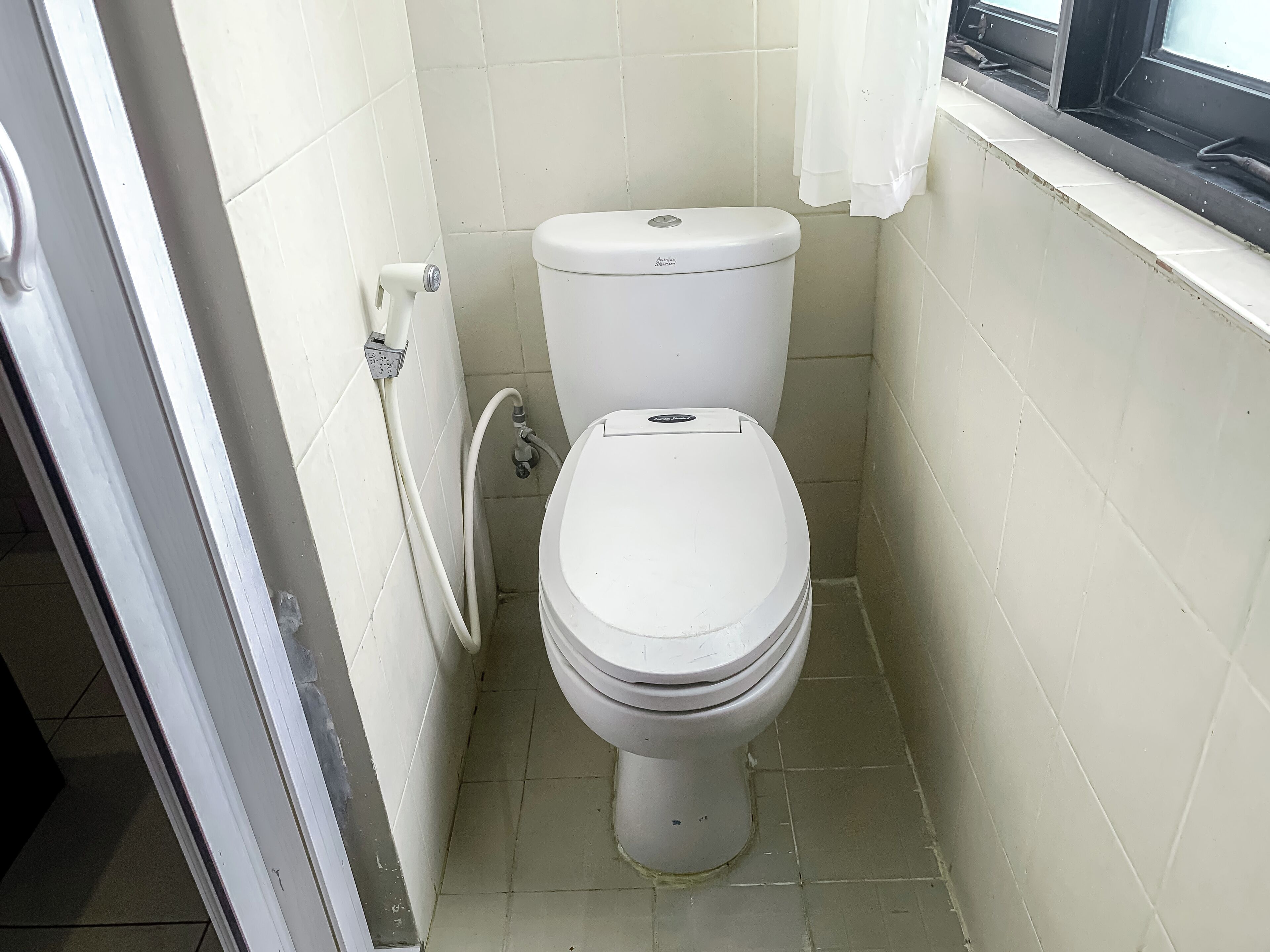 A small, clean bathroom with a toilet, a showerhead, and a window with a curtain.