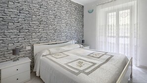 A bedroom with a stone wall, a bed with a patterned bedspread, a bedside table with a lamp, and a window with sheer curtains. - Apartment 'Da Ercolino' with Sea View, Balcony and Wi-Fi (Pitelli)