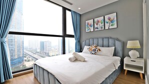 A modern hotel room with a large bed, a nightstand, a lamp, and a city view through the window. - Mira Housing- Vinhome Skylake Luxury Apartment  (Hanoi)