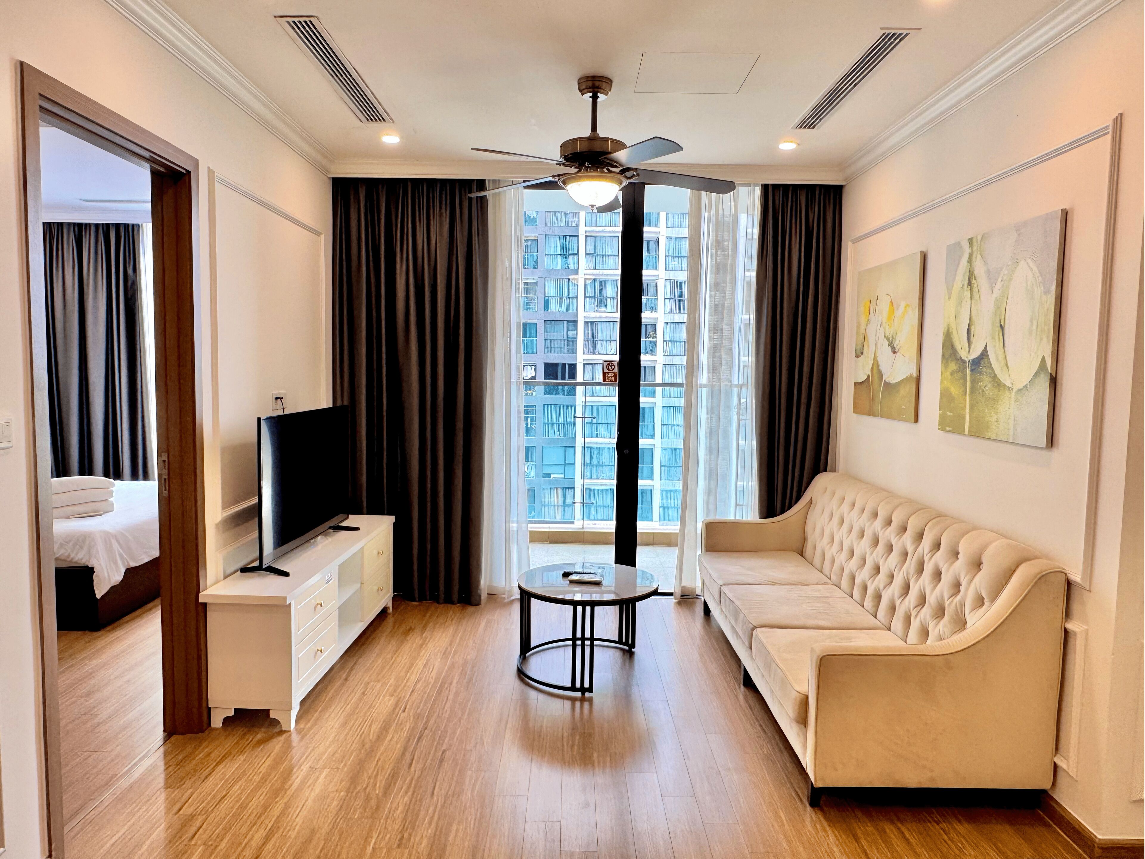 A modern hotel room with a flat-screen TV, a tufted sofa, a small round table, and a ceiling fan.