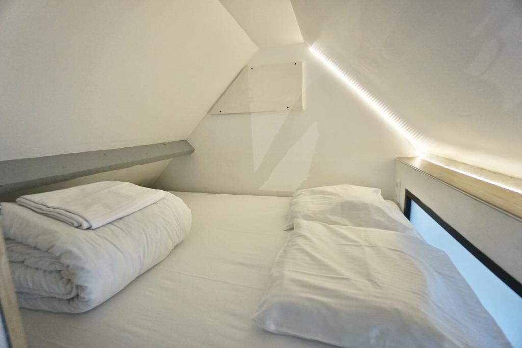 A small, single-bed room with a sloped ceiling, a bedside table, and a wall-mounted light fixture.