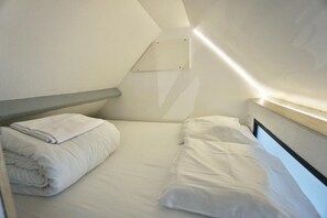 A small, single-bed room with a sloped ceiling, a bedside table, and a wall-mounted light fixture.
