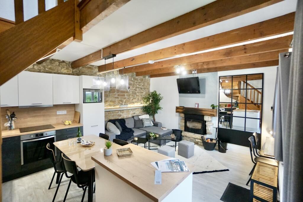 Una cocina moderna con pared de piedra, vigas de madera, zona de comedor con una mesa larga y sillas, sala de estar con sofá y chimenea, y una escalera que conduce a un nivel superior.