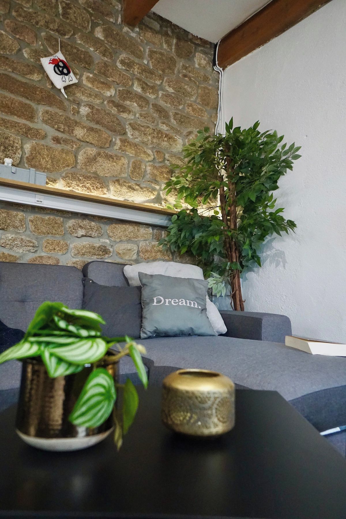 A cozy room with a stone wall, a grey sofa with a cushion labeled 'Dream.', a small plant on a table, and a decorative item hanging on the wall.