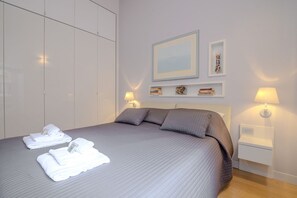 A neatly made bed with pillows, a bedside table with a lamp, and a wall-mounted shelf with books and decorative items.