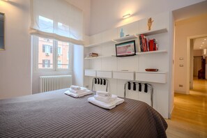 A hotel room with a bed, a window, a radiator, a shelf with books, and a television.