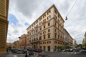 Un edificio de varios pisos con estilo arquitectónico clásico, que presenta una entrada en esquina y un nivel inferior visible con zona de estacionamiento.