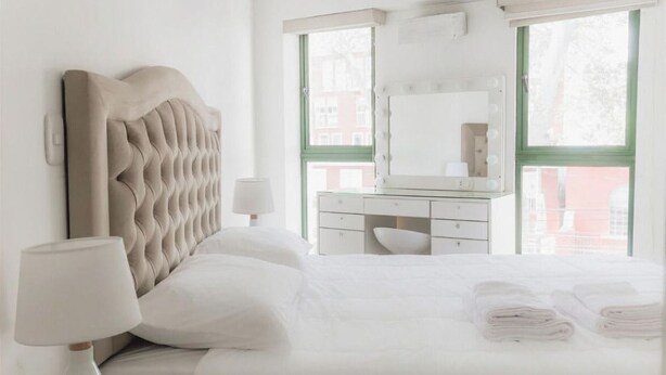 A bedroom with a tufted headboard, a bedside lamp, a dresser, a mirror, and a window with a view of buildings. - Elegance in Barranco - Cozy 2BR (Barranco)