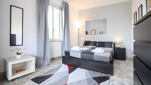 Un dormitorio con cama, mesitas de noche, cómoda, espejo, ventana con cortinas y una alfombra estampada.