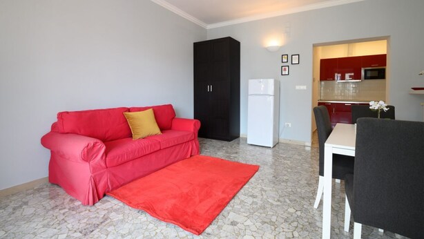 Un espacio habitable compacto con un sofá rojo, una pequeña mesa de comedor con sillas, una zona de cocina con armarios rojos y un refrigerador blanco.