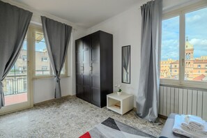 Una habitación con un armario grande, una mesita de noche pequeña con una planta, vistas a edificios por la ventana y un suelo de baldosas.