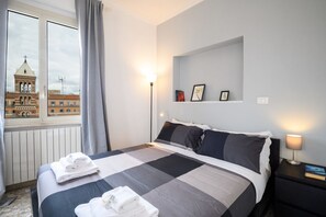 A hotel room with a bed, a nightstand, a lamp, a window with curtains, and a view of a building with a tower.