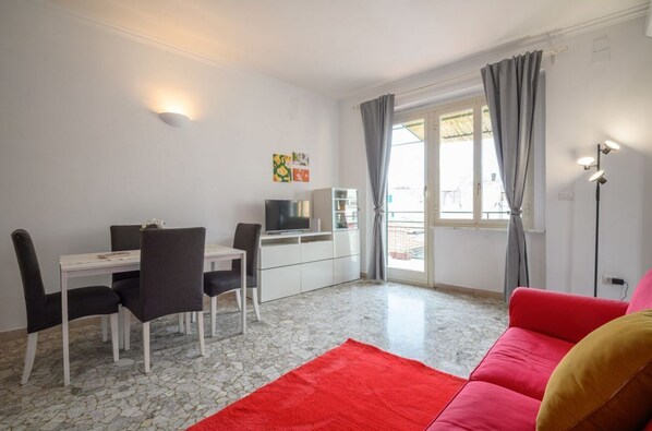 Una habitación con una mesa de comedor, sillas, televisor, sofá y una ventana con cortinas.