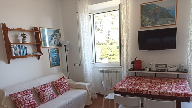 A cozy room with a white sofa, a dining table with chairs, a TV mounted on the wall, and a window with a view of buildings and greenery. - Stunning Sea View Apartment in the Heart of Riomaggiore (Riomaggiore)