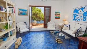 A living room with a white sofa, a coffee table, and a bookshelf. There is a door leading to an outdoor area with a view of trees and a building. - Villa Majus - with swimming pool and direct access to the sea in marine protected area  (BAGHERIA)