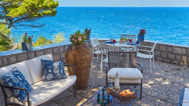 An outdoor seating area with a view of the sea, featuring a wicker chair, a white sofa, and a metal table with decorative items. - Villa Majus - with swimming pool and direct access to the sea in marine protected area  (BAGHERIA)