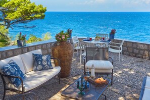An outdoor seating area with a view of the sea, featuring a wicker chair, a white sofa, and a metal table with decorative items.