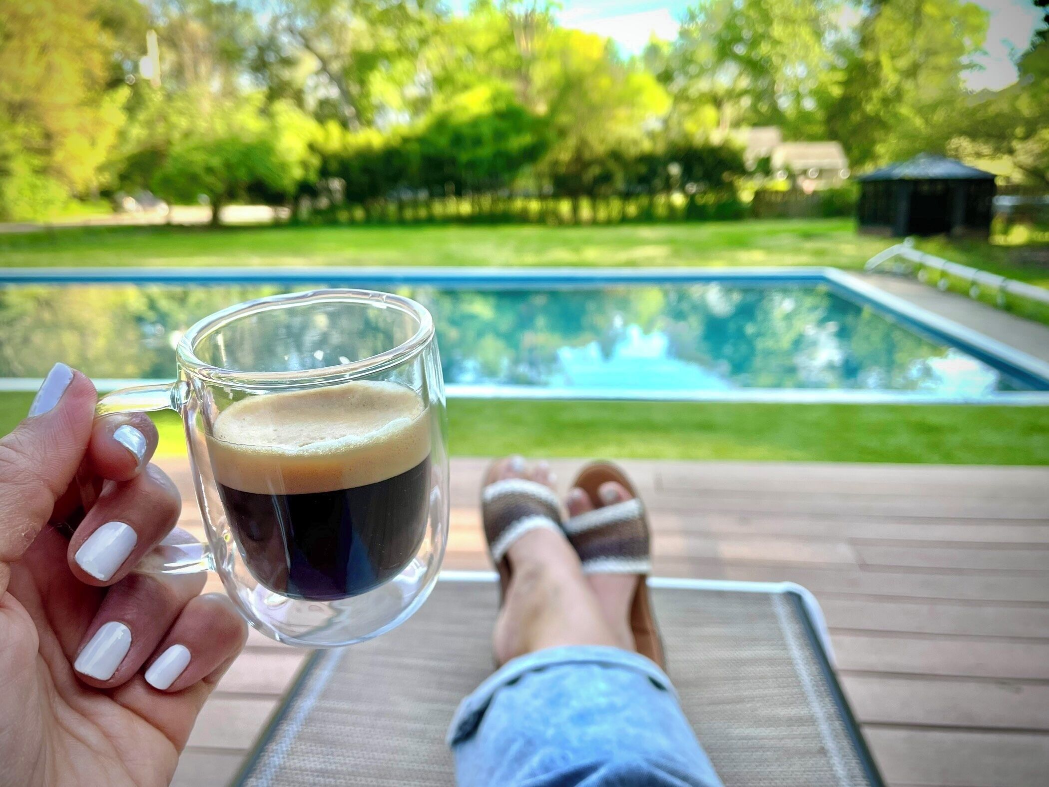 A person holding a glass of dark liquid, likely coffee, on a deck overlooking a swimming pool.