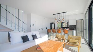 Un salon moderne avec un canapé, une table basse et des fauteuils en osier. On aperçoit à l’arrière-plan une cuisine avec un coin repas et une grande fenêtre.