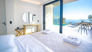 Une chambre d’hôtel moderne avec un grand lit, un miroir rond, une petite table et une vue sur la mer par une porte vitrée.