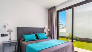 A modern hotel room with a large bed, two bedside lamps, a nightstand, and a view of a green lawn through a sliding glass door.