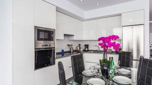 A modern kitchen with white cabinets, a built-in microwave, oven, and a refrigerator. There is a dining table with chairs, a vase of flowers, and a glass top.