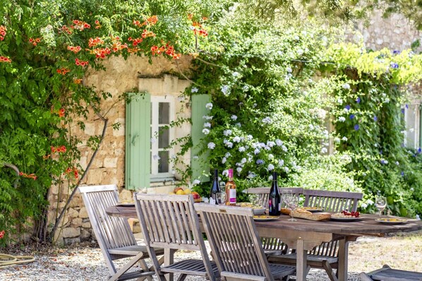 An outdoor dining area with a rustic wooden table and chairs, surrounded by lush greenery and flowering plants.