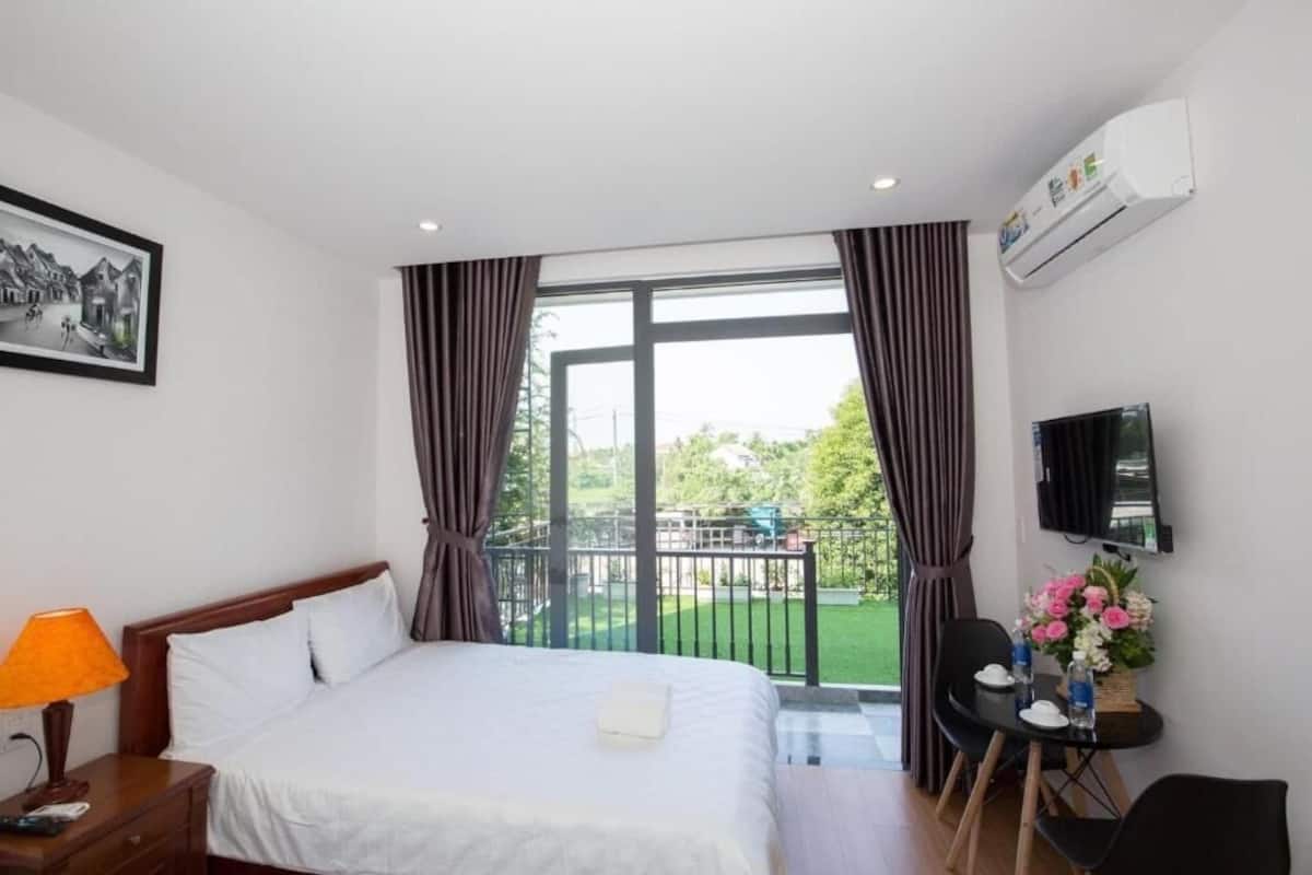 A hotel room with a bed, a desk with a chair, a TV, and a balcony with curtains.