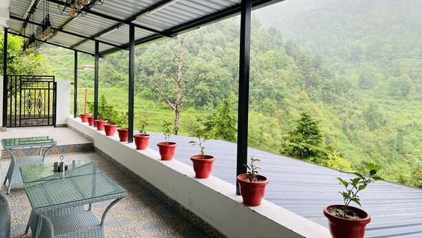 A covered balcony with a glass table and chairs, potted plants, and a view of a lush green valley. - StudioZ Paradise Hills Mussoorie (Mussoorie)