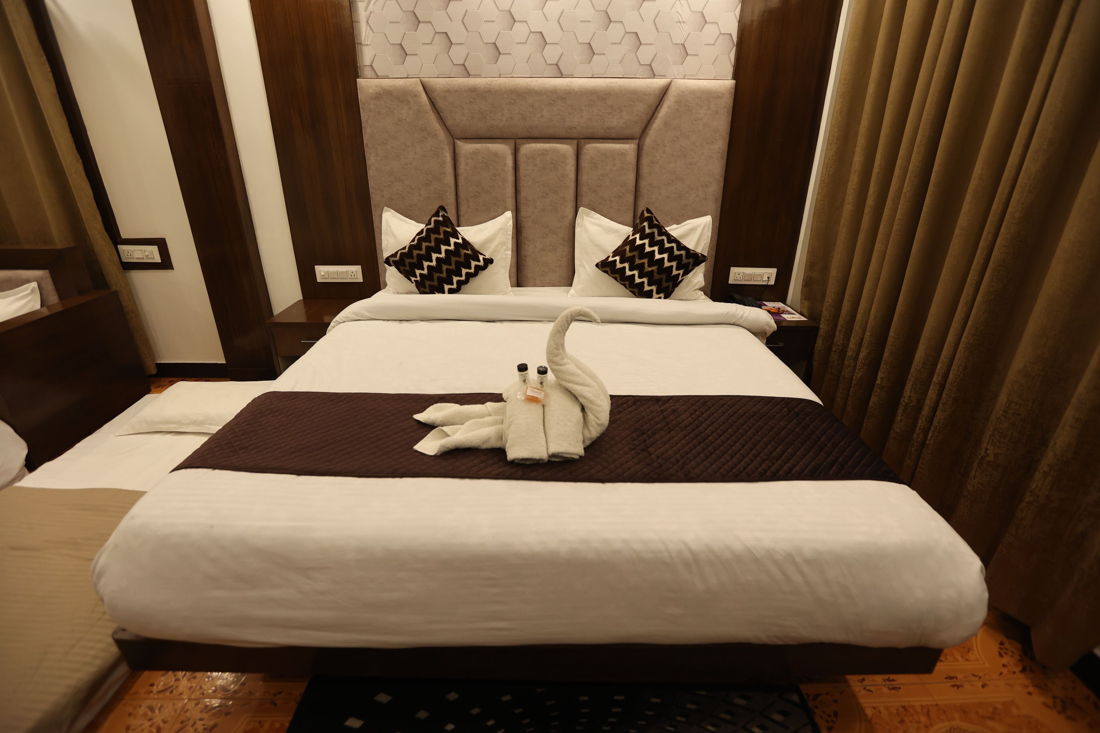A hotel room with a large bed, two pillows, a headboard, a nightstand, and a curtain.