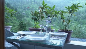 A dining table set with plates, cutlery, and glasses, surrounded by potted plants, on a balcony with a view of a forested landscape. - StudioZ Paradise Hills Mussoorie (Mussoorie)