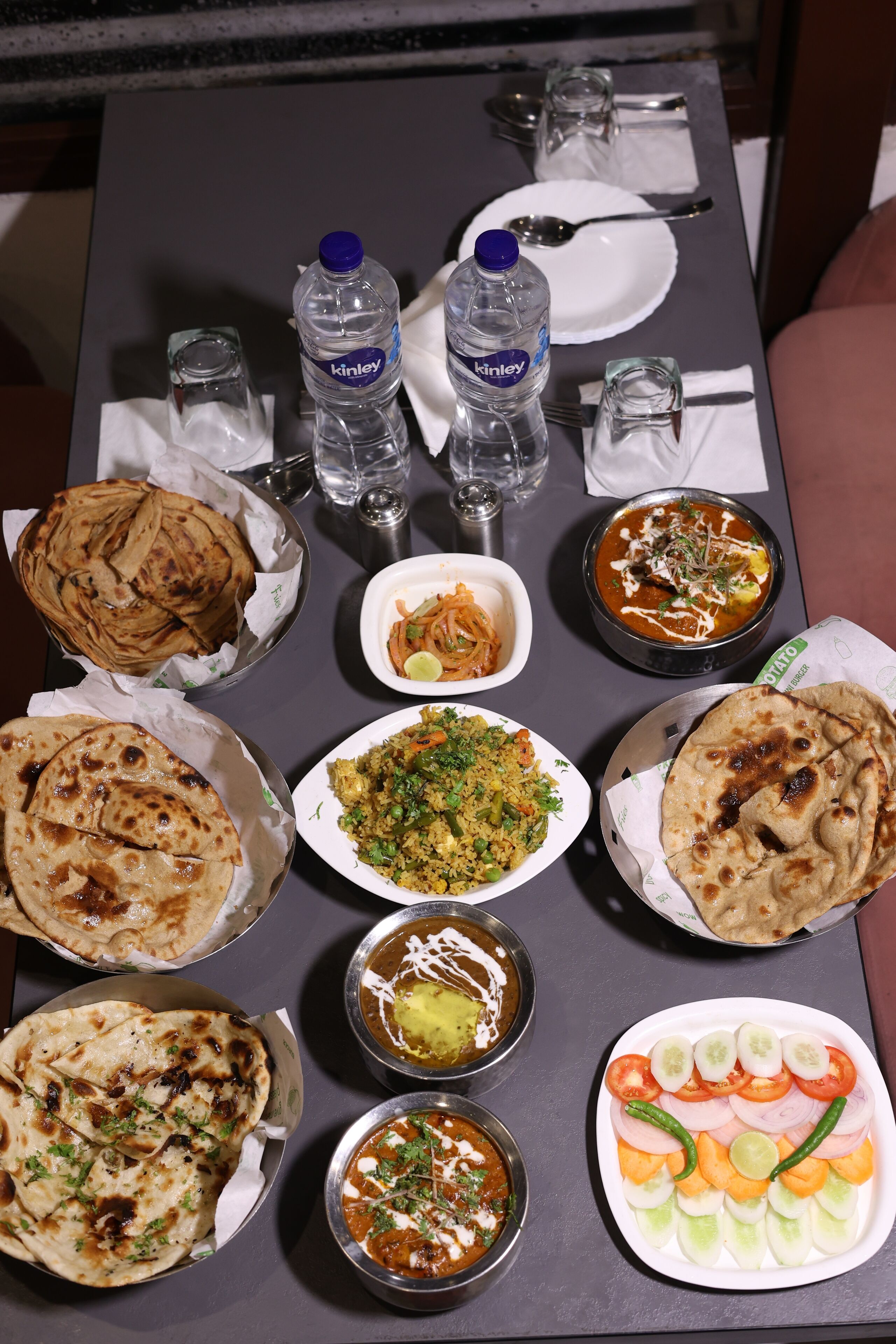 A table with various dishes, including naan bread, curry, and a salad. There are two bottles of water and a plate with cutlery.