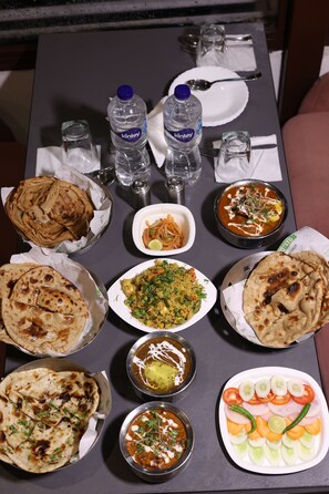 A table with various dishes, including naan bread, curry, and a salad. There are two bottles of water and a plate with cutlery. - StudioZ Paradise Hills Mussoorie (Mussoorie)