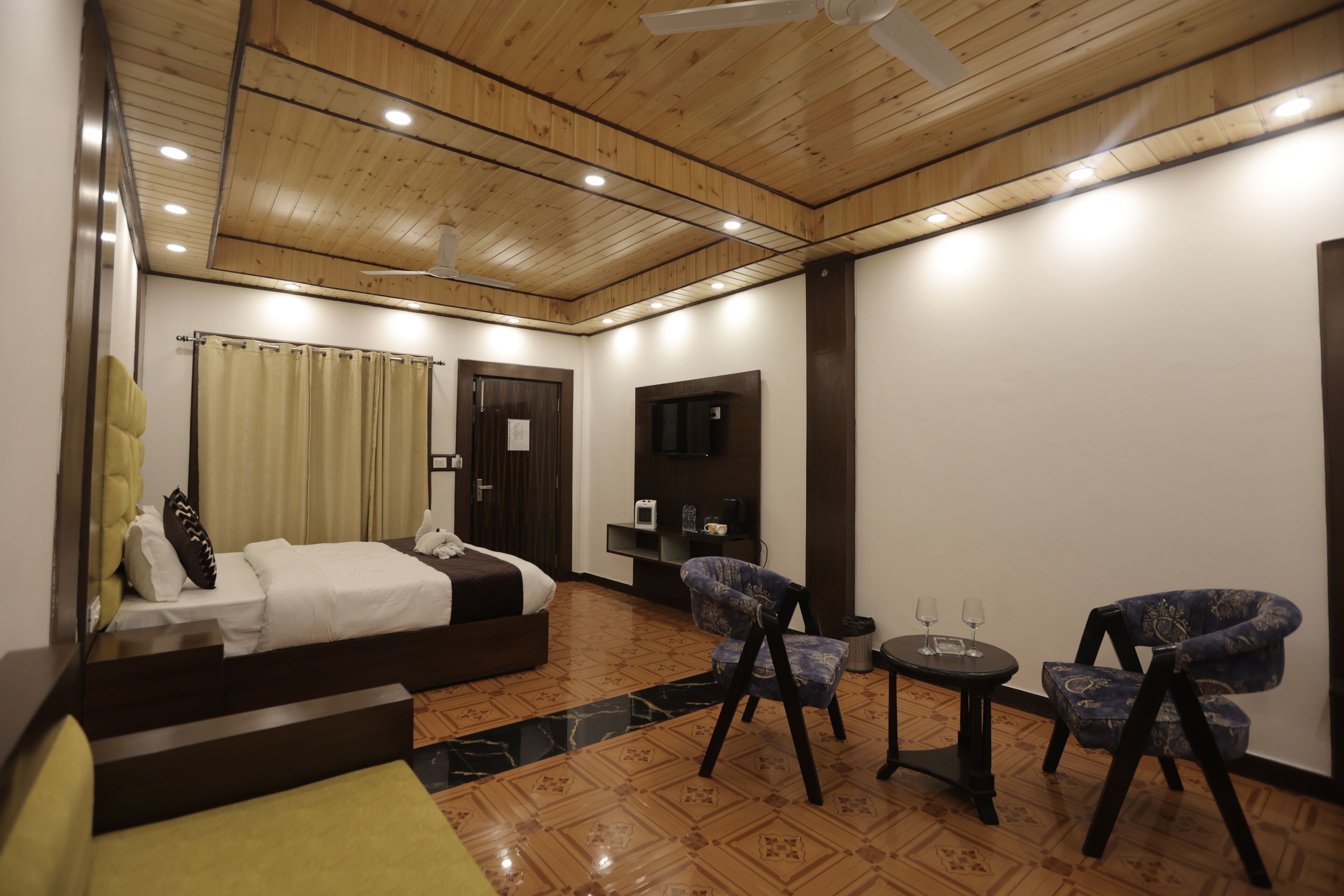 A hotel room with a bed, two chairs, a small table, a TV, and a ceiling fan.