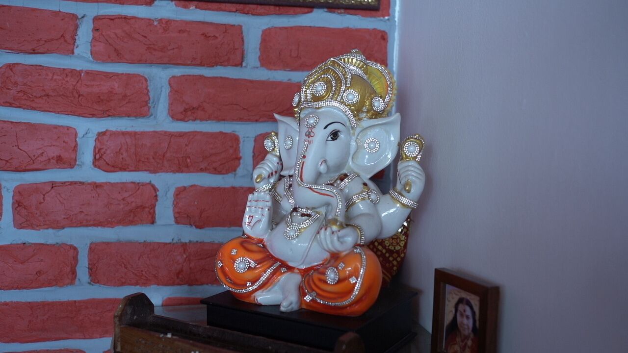 Una estatua de una deidad con corona y joyas, sobre un pedestal de madera frente a un muro de ladrillo.