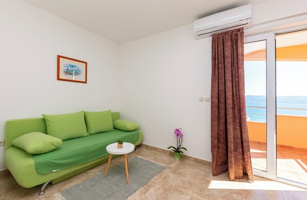 A green sofa with a small table and a potted plant in a room with a view of the sea through a sliding door.