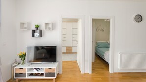 A modern, well-lit room with a flat-screen TV on a white TV stand, a small plant, and a clock on the wall. - Apartment L and I (Sibenik)